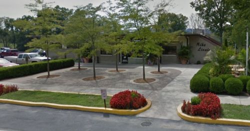 A landscaped area with trees and flower beds in front of a building, featuring a parking lot and a clear sky.