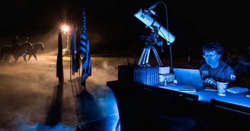 A person works on a laptop at night, with a telescope and flags nearby, while horses are seen in the background.