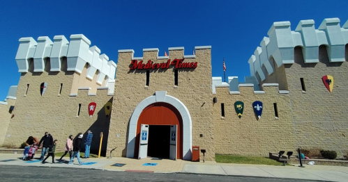 Exterior of a Medieval Times castle with flags, featuring a large entrance and visitors walking in front.