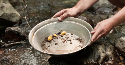 A person holds a pan with gravel and gold nuggets over a stream, surrounded by rocks and flowing water.