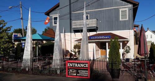 A European restaurant with a fenced outdoor seating area, featuring tall, pointed sculptures and a welcoming entrance sign.