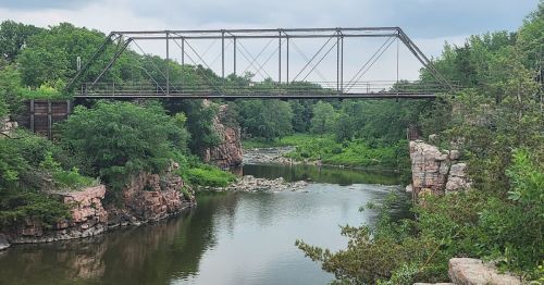 A historic metal bridge spans a river surrounded by lush greenery and rocky cliffs under a cloudy sky.