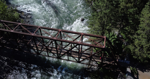 Aerial view of a rusted metal bridge over a rushing river, surrounded by lush green trees.