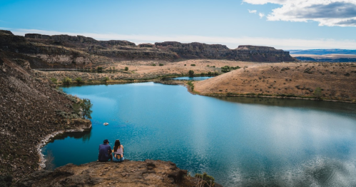 A couple sits on a rocky ledge overlooking a serene lake surrounded by hills and blue skies.