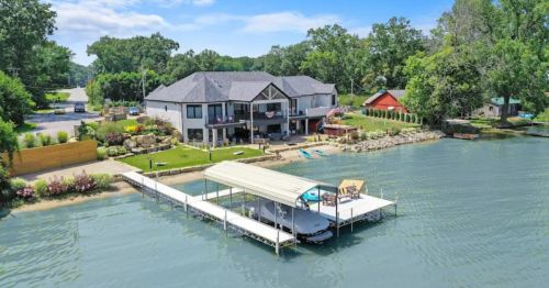 A large lakeside house with a dock, surrounded by greenery and a clear blue sky.