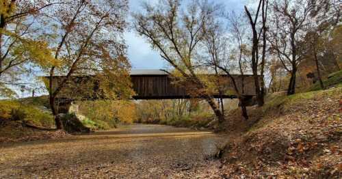 A covered bridge spans a dry riverbed, surrounded by autumn trees with colorful leaves.