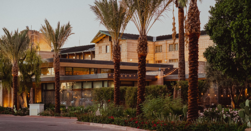 A modern building surrounded by palm trees and lush greenery, illuminated by warm lights at dusk.
