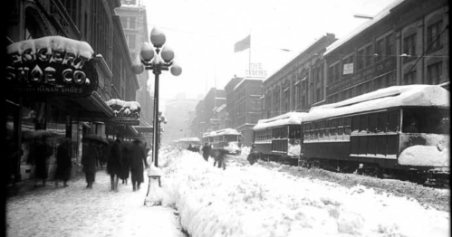 A snowy street scene with vintage streetcars and pedestrians in heavy snow, buildings in the background.