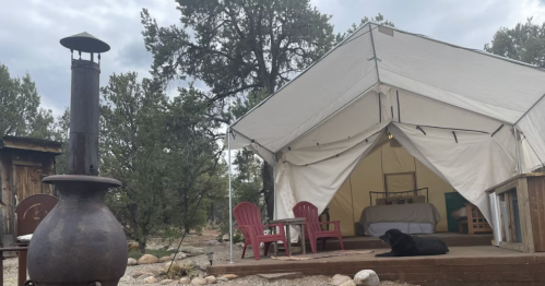 A cozy tent with a bed and chairs, surrounded by trees, with a black dog resting on the porch. A chiminea stands nearby.