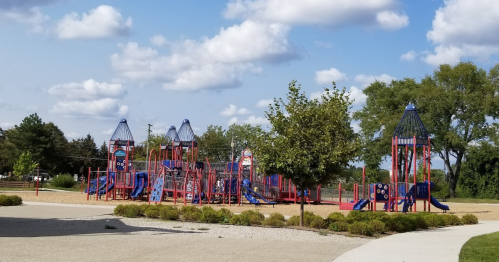 Colorful playground with slides and climbing structures, surrounded by trees and a blue sky with fluffy clouds.