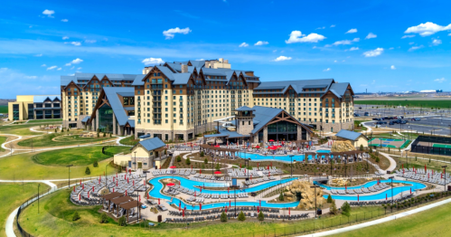 A large hotel with a pool area, surrounded by green lawns and blue skies, featuring modern architecture and outdoor seating.