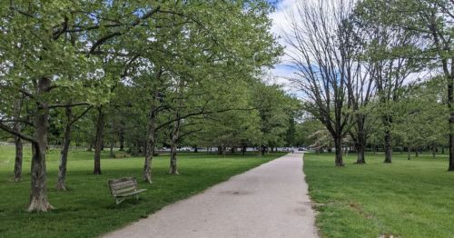 A peaceful park scene featuring a paved path lined with trees and a bench, under a partly cloudy sky.