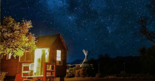 A cozy cabin and a teepee under a starry night sky, with warm light glowing from the cabin's open door.