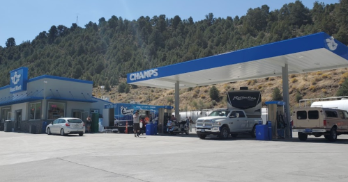 A gas station with a convenience store, surrounded by trees and mountains, with vehicles refueling at the pumps.