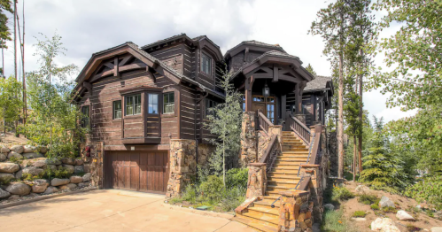 A large, rustic wooden house with stone accents, surrounded by trees and featuring a staircase leading to the entrance.