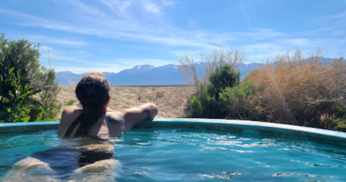 A person relaxes in a hot tub, gazing at a scenic view of mountains and a clear blue sky.