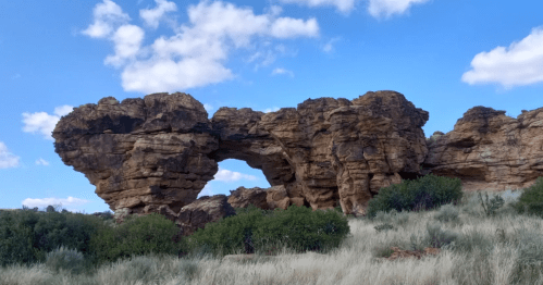 A natural rock formation with an arch, surrounded by green shrubs and a blue sky with scattered clouds.