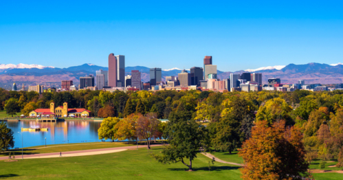 A vibrant city skyline with mountains in the background, surrounded by a park and a lake on a clear day.