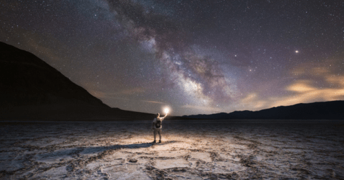 An astronaut stands on a barren landscape, holding a light, under a starry sky with the Milky Way visible.