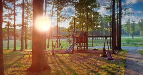 A playground with swings and slides, surrounded by trees, illuminated by the warm glow of the setting sun.
