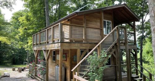 A two-story wooden cabin surrounded by trees, featuring a balcony and stairs leading to the entrance.