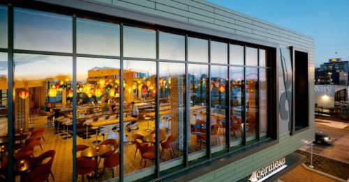 A modern restaurant with large windows, colorful pendant lights, and a view of the city skyline at sunset.