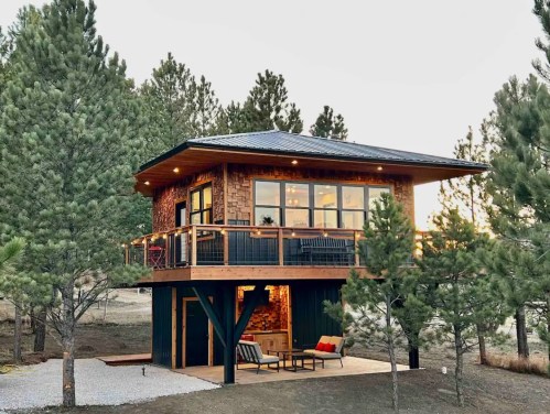 A modern cabin with a wooden exterior, elevated on stilts, surrounded by pine trees and featuring a cozy outdoor seating area.