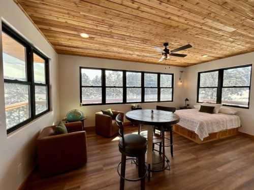 Cozy room with wooden ceiling, large windows, a bed, seating area, and a round table, surrounded by nature.