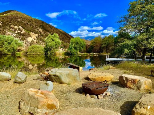A serene lakeside scene with a fire pit, surrounded by rocks and lush greenery, under a blue sky with fluffy clouds.