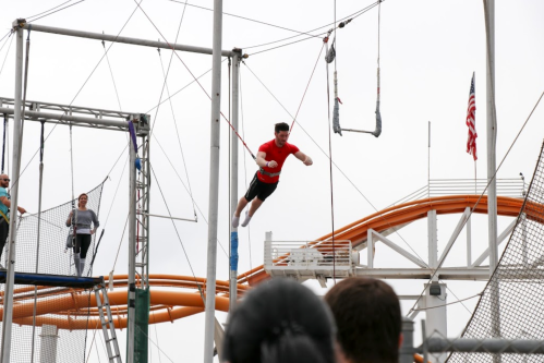 A person in a red shirt swings on a trapeze, with spectators and a roller coaster in the background.