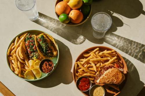 Two plates of food: one with tacos and fries, the other with a sandwich and fries, alongside a bowl of fresh fruit.
