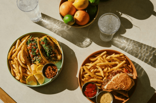 Two plates of food: one with tacos and fries, the other with a sandwich and fries, alongside a bowl of fresh fruit.