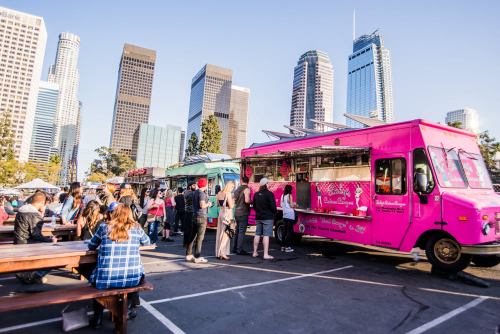A vibrant food truck event in an urban setting, with people enjoying food and tall buildings in the background.
