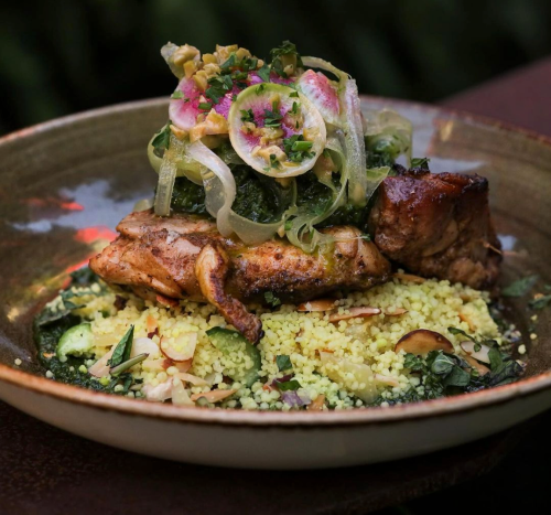 A beautifully plated dish featuring grilled chicken atop couscous, garnished with herbs, pickled vegetables, and onions.