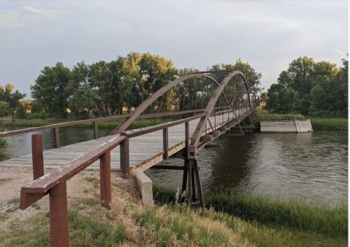 A wooden arch bridge spans a calm river, surrounded by green trees and grassy banks under a cloudy sky.