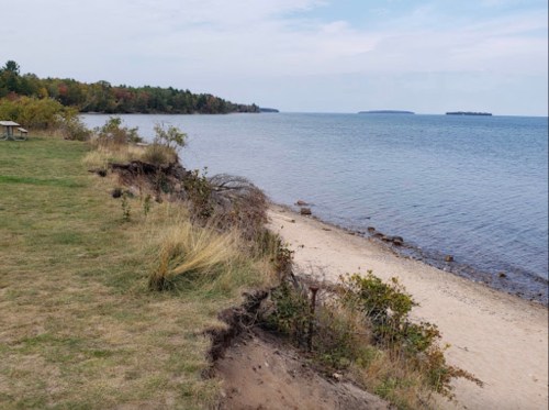 A serene lakeside view with sandy shore, grassy area, and distant islands under a cloudy sky.