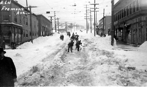 Historic black-and-white photo of a snowy street with people walking and snow piled high on either side.