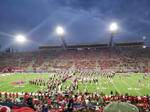 A large crowd at a stadium during a halftime show, with performers in red and white uniforms on the field.