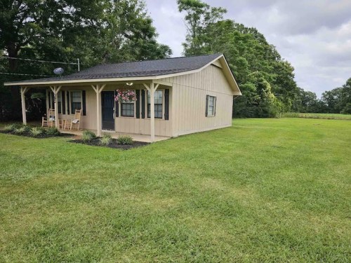 A small, light-colored house with a porch, surrounded by green grass and trees under a cloudy sky.