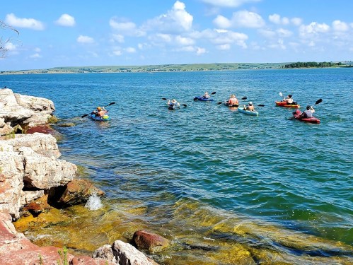 A group of kayakers paddles on a calm lake, with rocky shores and blue skies in the background.