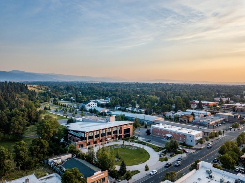 Aerial view of a town with green spaces, buildings, and mountains in the background under a clear sky at sunset.