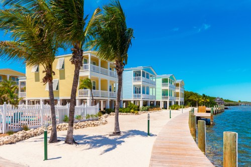 Colorful beachside buildings with palm trees along a wooden walkway by the water under a clear blue sky.
