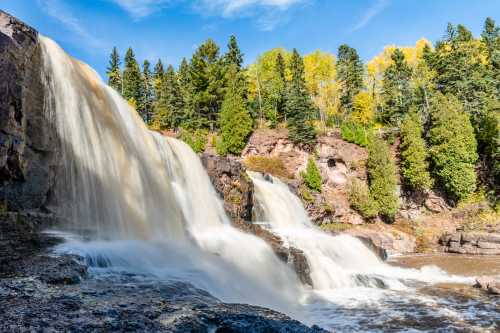 A scenic waterfall cascading over rocks, surrounded by lush green trees and autumn foliage under a clear blue sky.