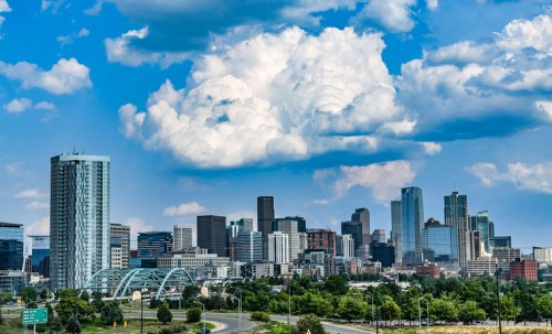 A vibrant city skyline with tall buildings under a blue sky filled with fluffy clouds. Green trees in the foreground.