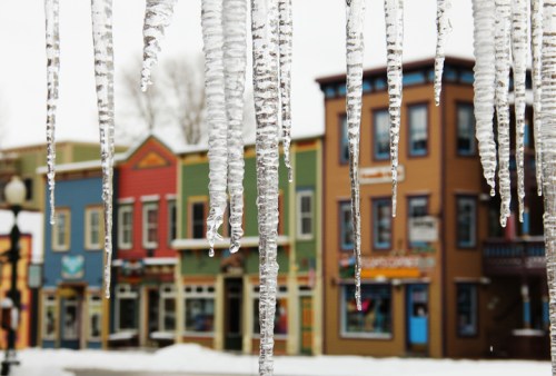 Icicles hang in the foreground, with colorful buildings blurred in the background on a snowy day.