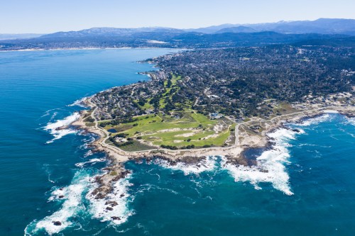Aerial view of a coastal landscape with rocky shores, green parks, and distant mountains under a clear blue sky.