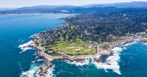Aerial view of a coastal area with lush greenery, rocky shores, and a clear blue ocean under a bright sky.