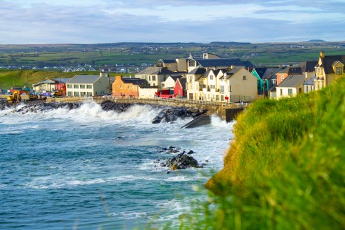 Coastal view of a small town with colorful buildings, waves crashing against the shore, and green hills in the background.