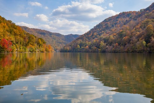 A serene lake surrounded by colorful autumn foliage and rolling hills under a partly cloudy sky.