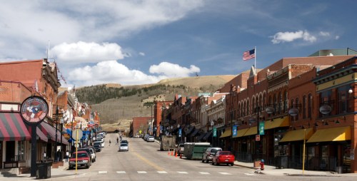 A charming street lined with historic buildings, shops, and parked cars, set against a backdrop of mountains and blue sky.